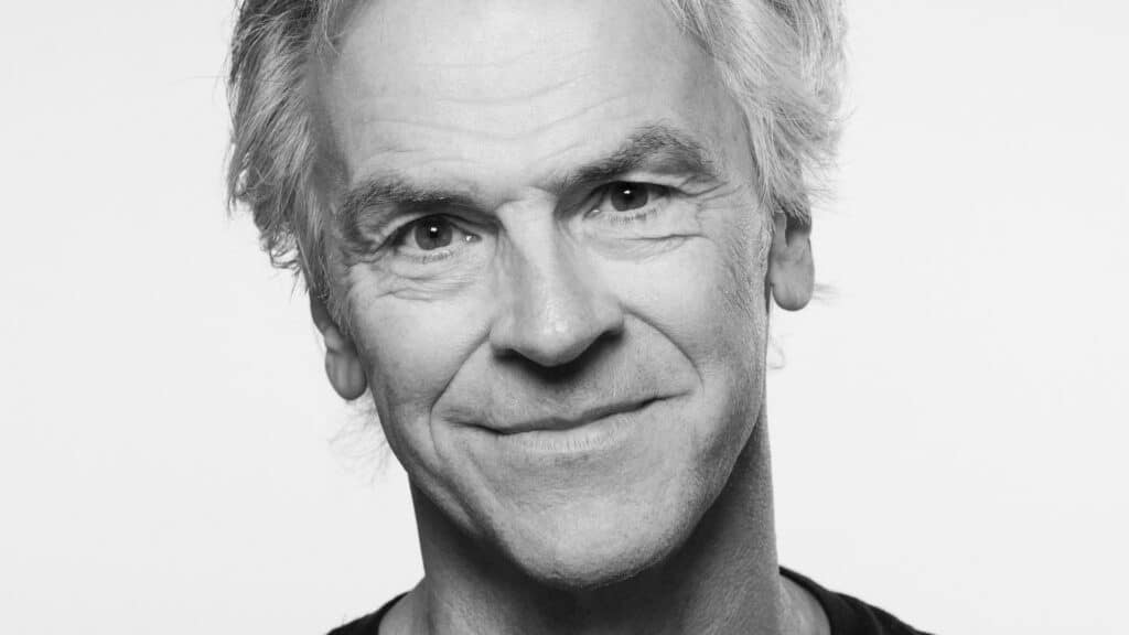 Et nærbilde i gråtoner av en eldre mann med grått hår, som smiler mykt og ser rett inn i kameraet mot en ensfarget bakgrunn.