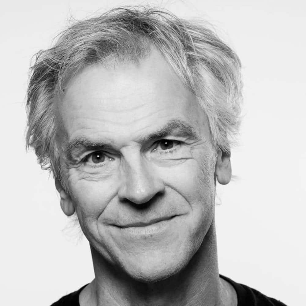 Svart-hvitt portrett av en smilende middelaldrende mann med ustelt hår.