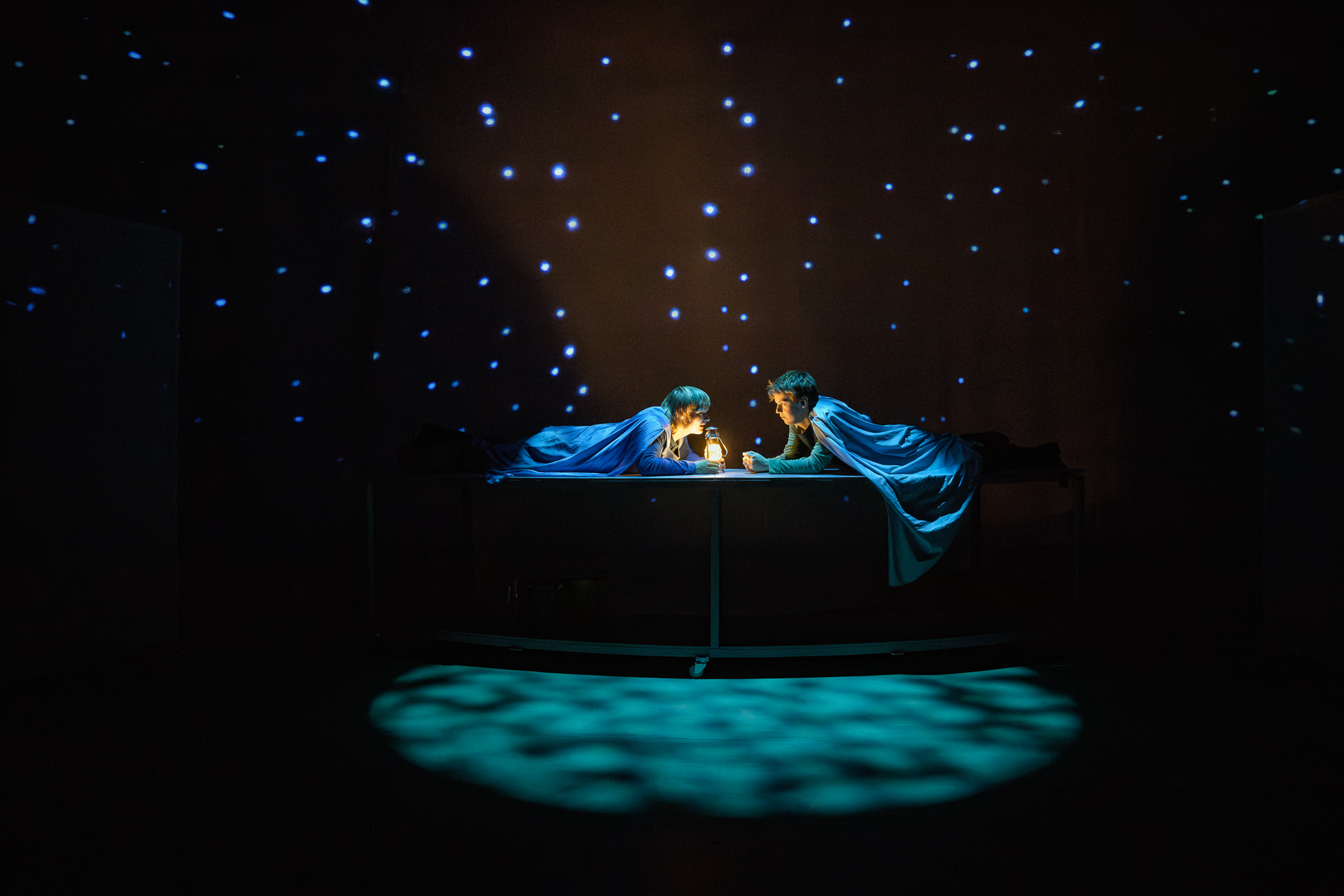 To personer i blå kapper ligger vendt mot hverandre over et bord, opplyst av en liten lampe, med en bakgrunn av projiserte stjerner og blå belysning på gulvet.