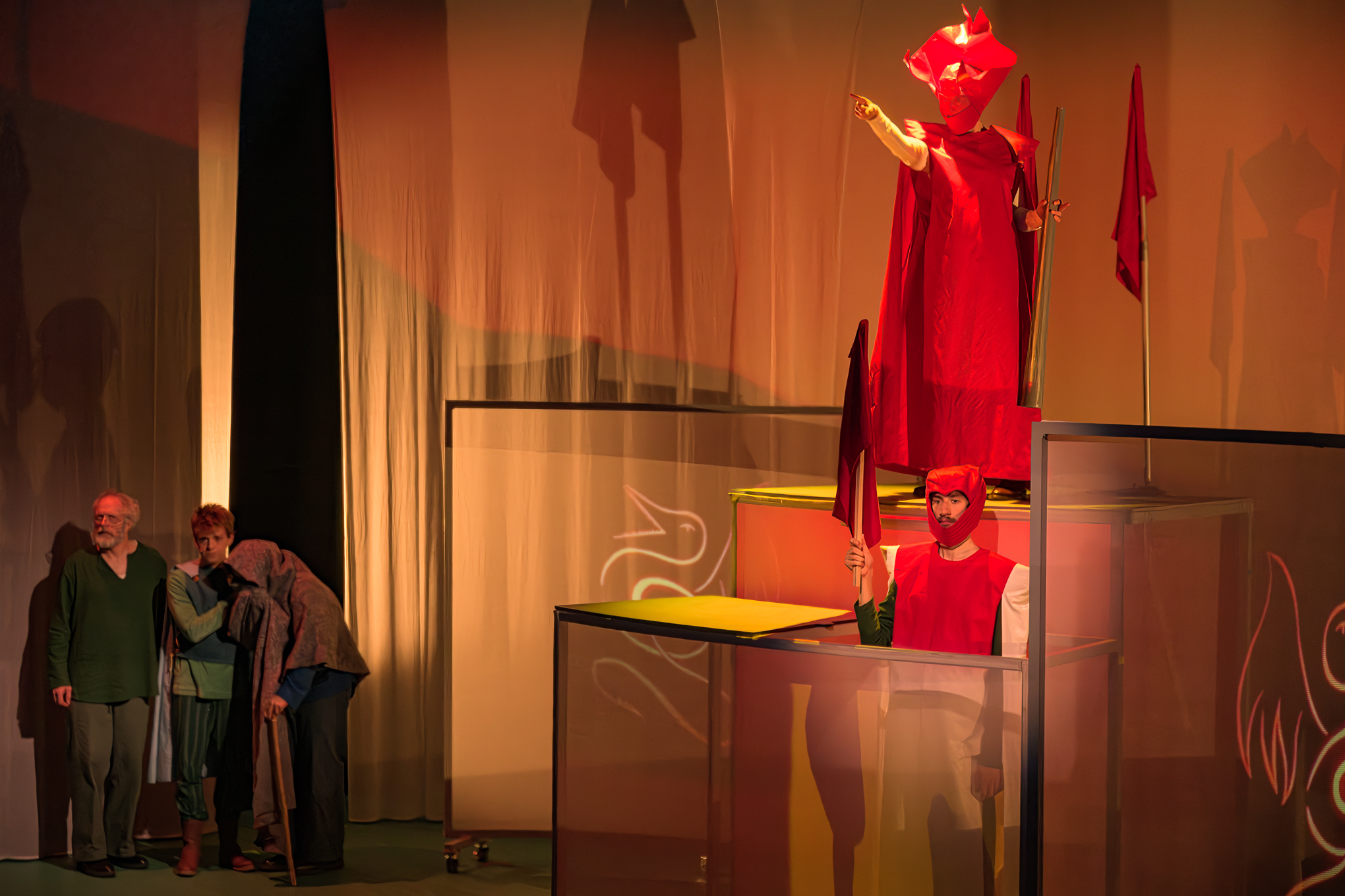 Tre personer står til venstre, mens to utøvere i røde kostymer holder flagg og poserer på opphøyde plattformer i en teatralsk scenografi med varm belysning og skygger.