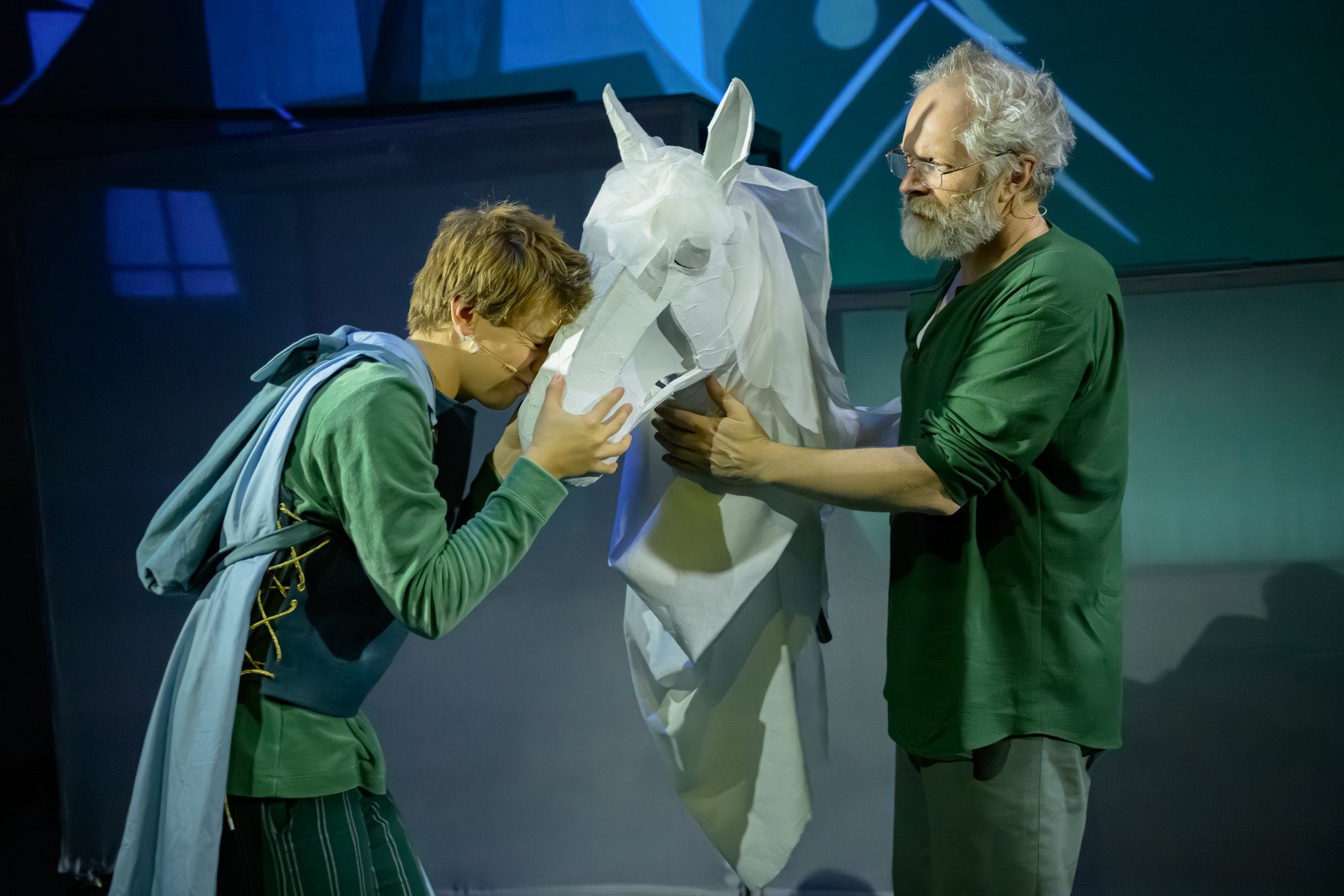To menn interagerer med en stor, hvit papirhestfigur på scenen; den ene holder hodet til figuren mens den andre lener seg tett inntil, begge iført grønne skjorter.