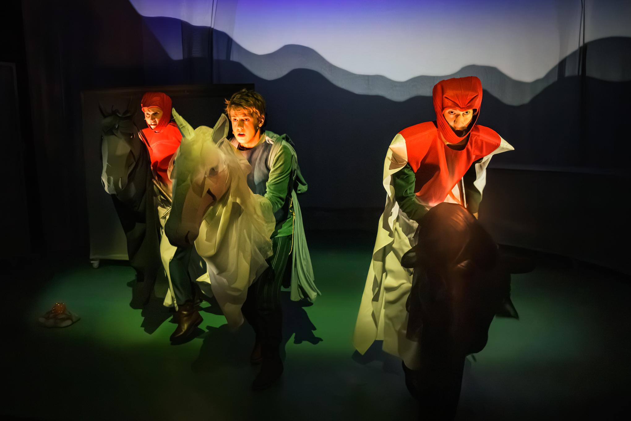 Tre skuespillere i fargerike kostymer og hodeplagg opptrer på scenen med hesterekvisitter i dramatisk lyssetting og et skyggefullt bakteppe.