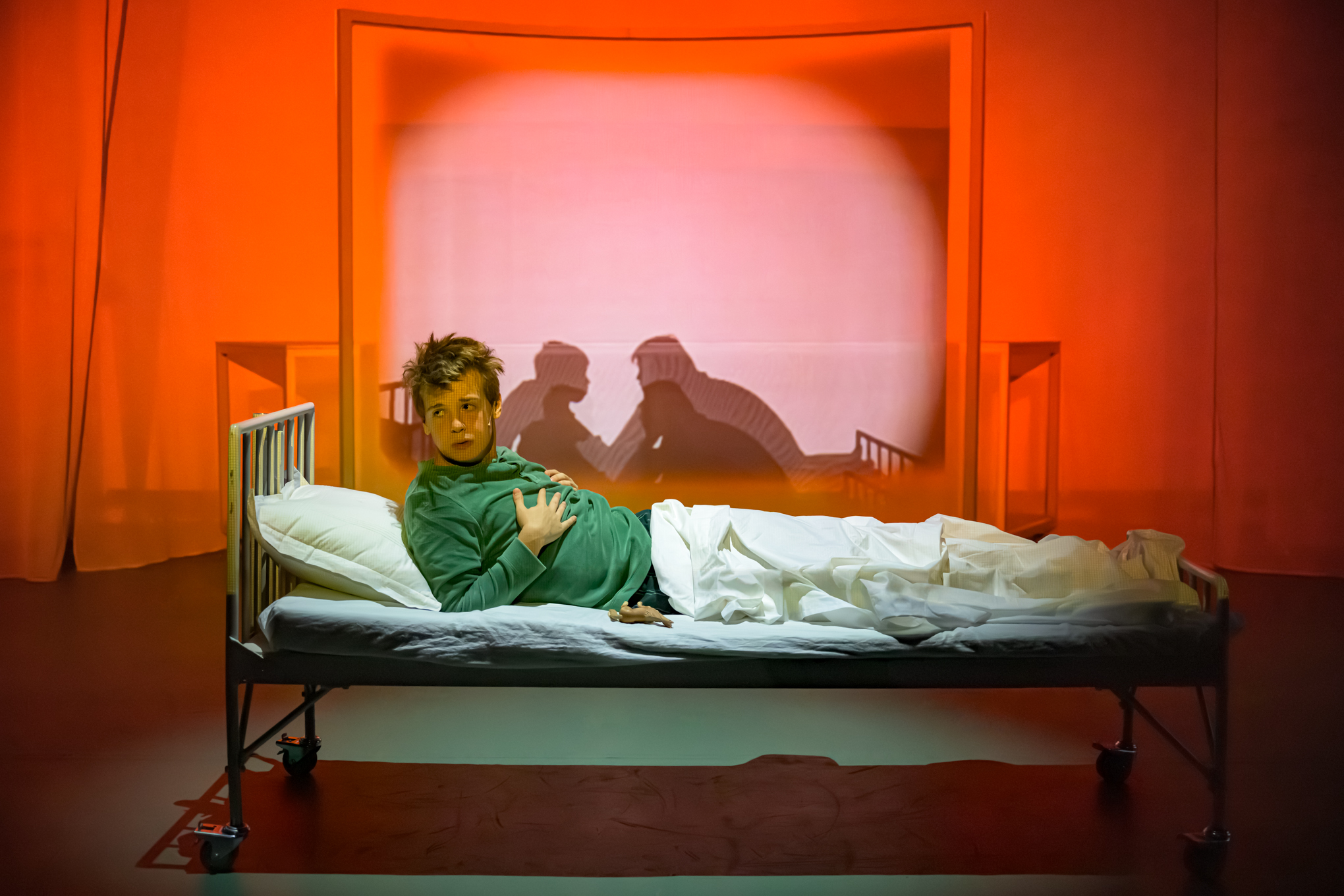 En person ligger i en sykehusseng på en scene med rød belysning som kaster skygger av to personer som holder hverandre i hendene på veggen bak dem.
