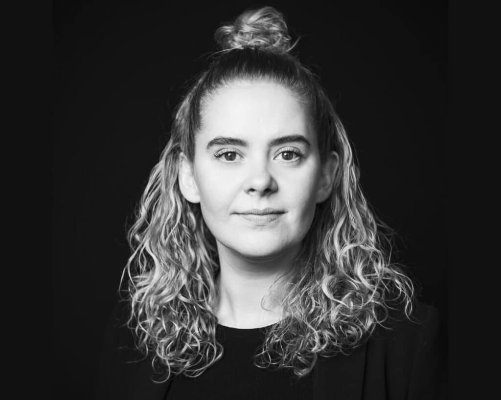 Portrettbilde av en kvinne med krøllete hår, satt opp i en halv oppsatt knute, mot en svart bakgrunn.
