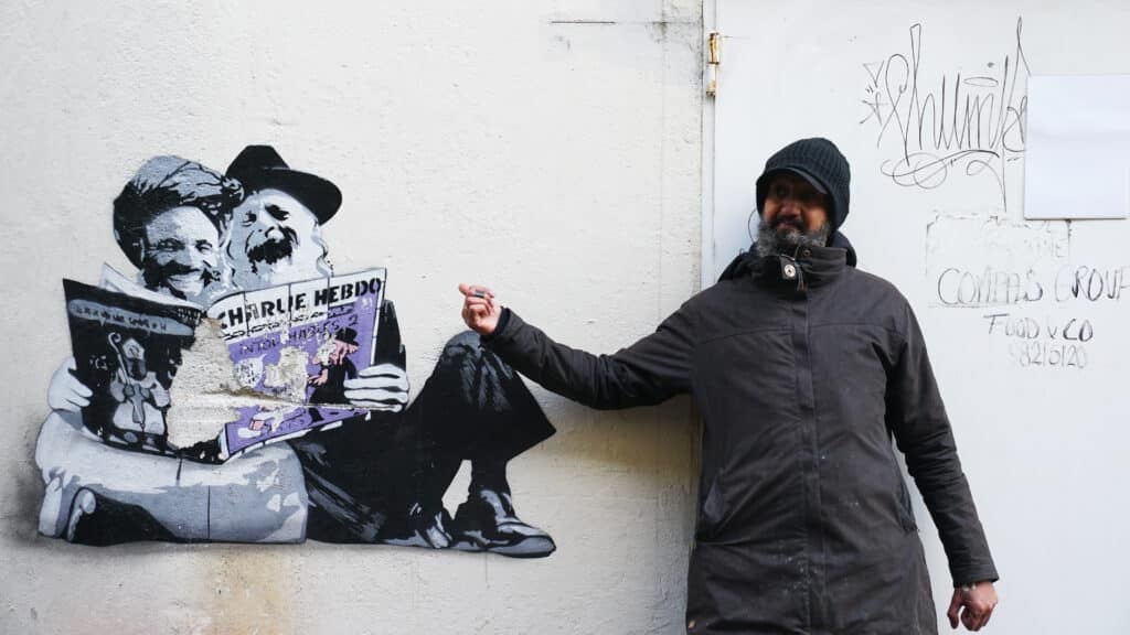 En mann i mørk frakk og lue står ved siden av en veggkunst av to figurer som leser et Charlie Hebdo-magasin. Veggen har også graffiti og en hvit dør.