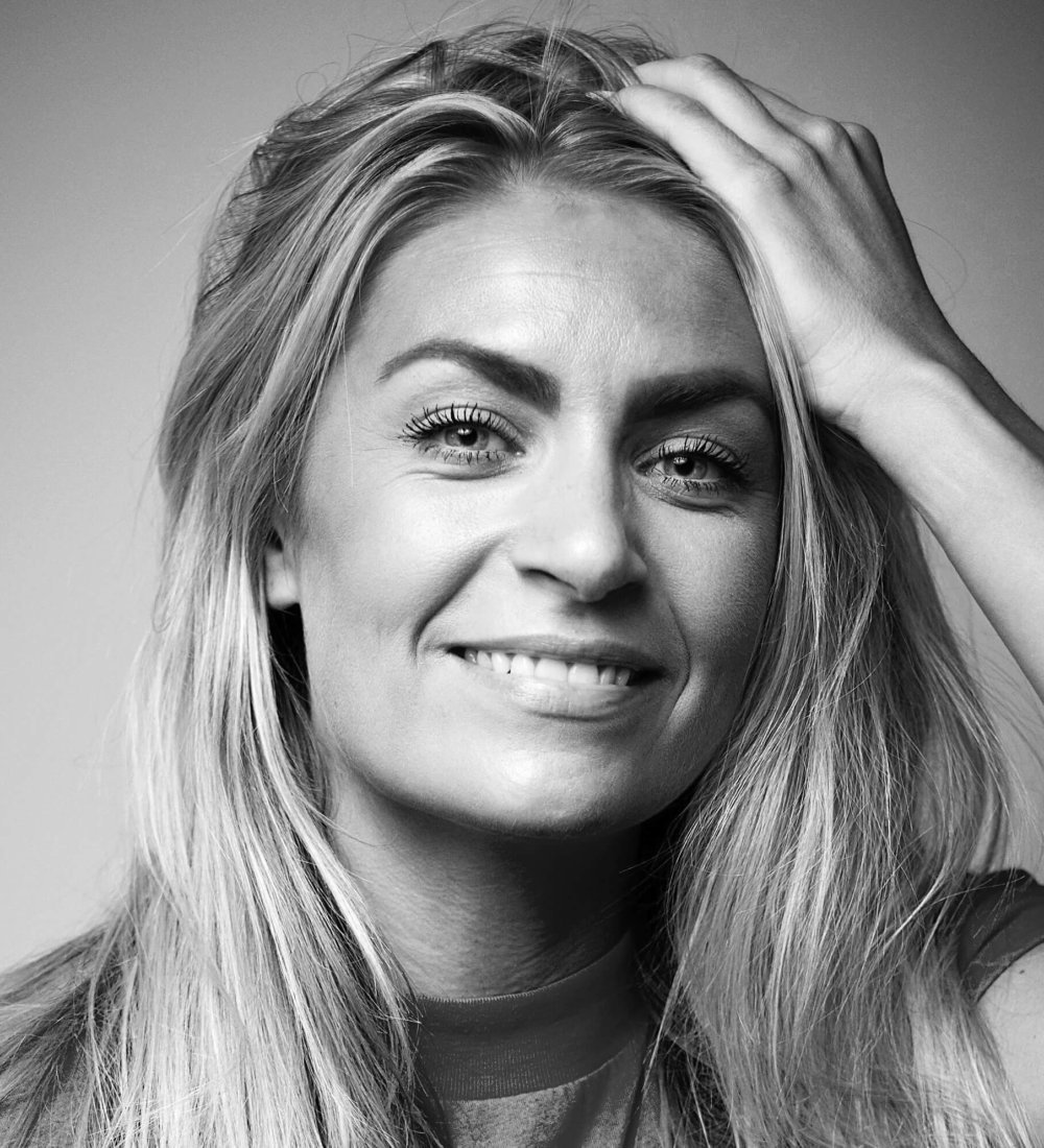 Elise Berg-Hansen_2 Et svart-hvitt portrett av en smilende kvinne med hånden i håret.