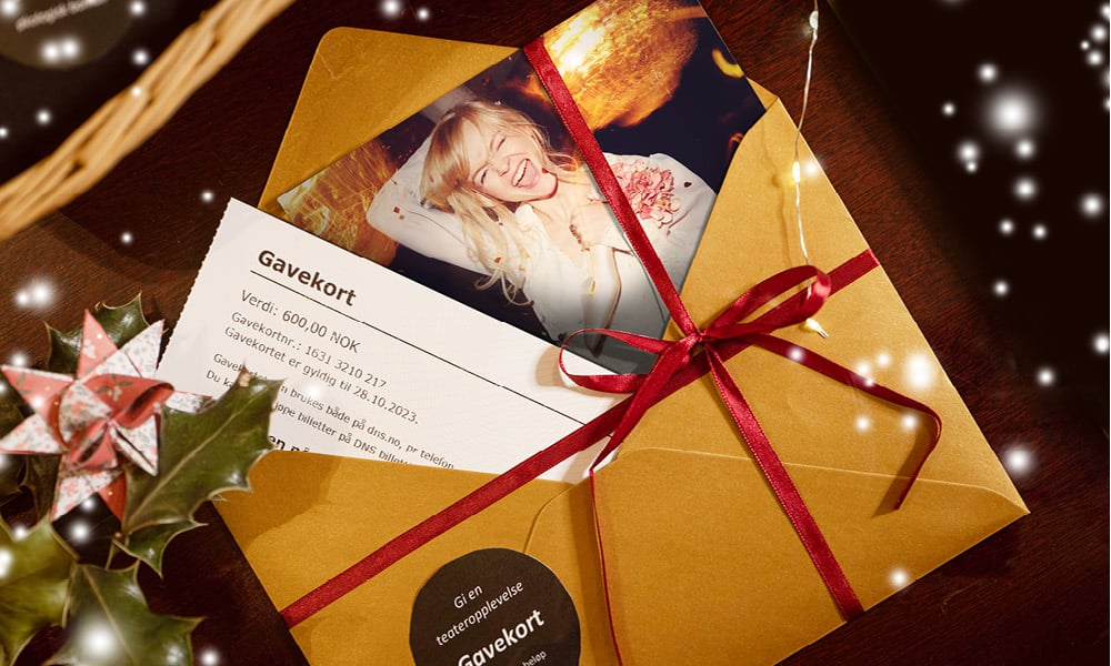 En brun konvolutt med rødt bånd inneholder et gavekort og et bilde av en smilende kvinne, og konvolutten er omkranset av festlig pynt og lys.