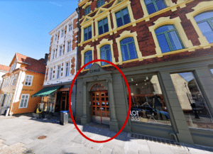 Gatebilde av et inngangsparti merket "CIMLE" med en dobbeltdør i tre, innrammet av søyler, og ved siden av butikkvinduer og en gul bygning.