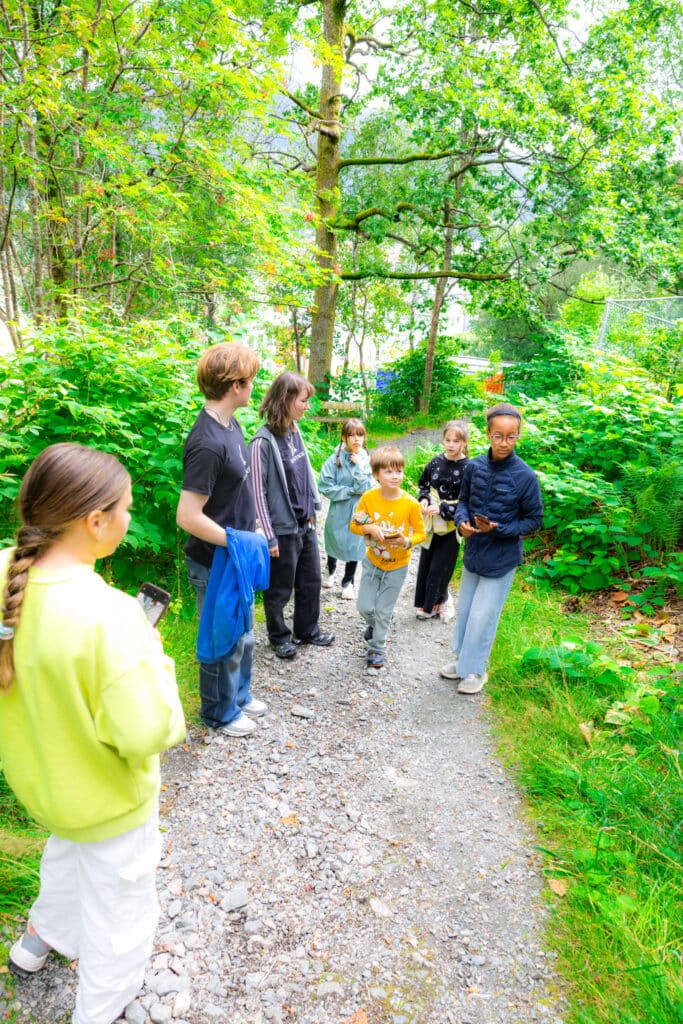 En gruppe barn og to voksne står og snakker sammen på en grussti omgitt av grønne trær og busker i et skogsområde.