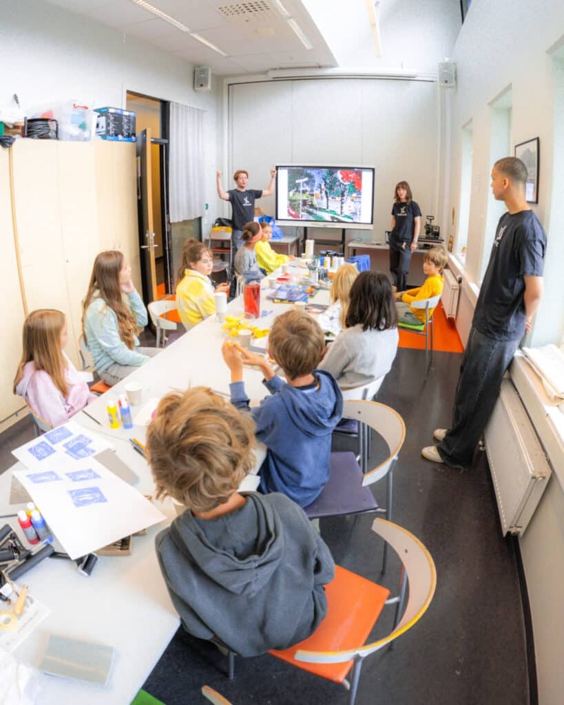 En gruppe barn sitter rundt et bord med kunst- og håndverksmaterialer, mens to voksne står foran i klasserommet ved siden av en stor skjerm som viser et bilde.
