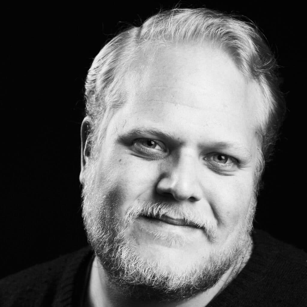 En skjeggete mann med kort, lyst hår og et svakt smil ser inn i kameraet mot en svart bakgrunn.