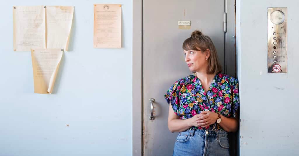 En kvinne i blomstrete bluse og jeans står ved en metalldør og ser til venstre. På veggen ved siden av er det montert papirer og et kontrollpanel for heisen.