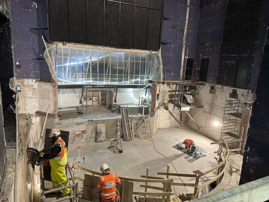 Bygningsarbeidere iført verneutstyr arbeider inne i en delvis revet bygning, mens stillaser, verktøy og bråte ligger spredt rundt på byggeplassen.