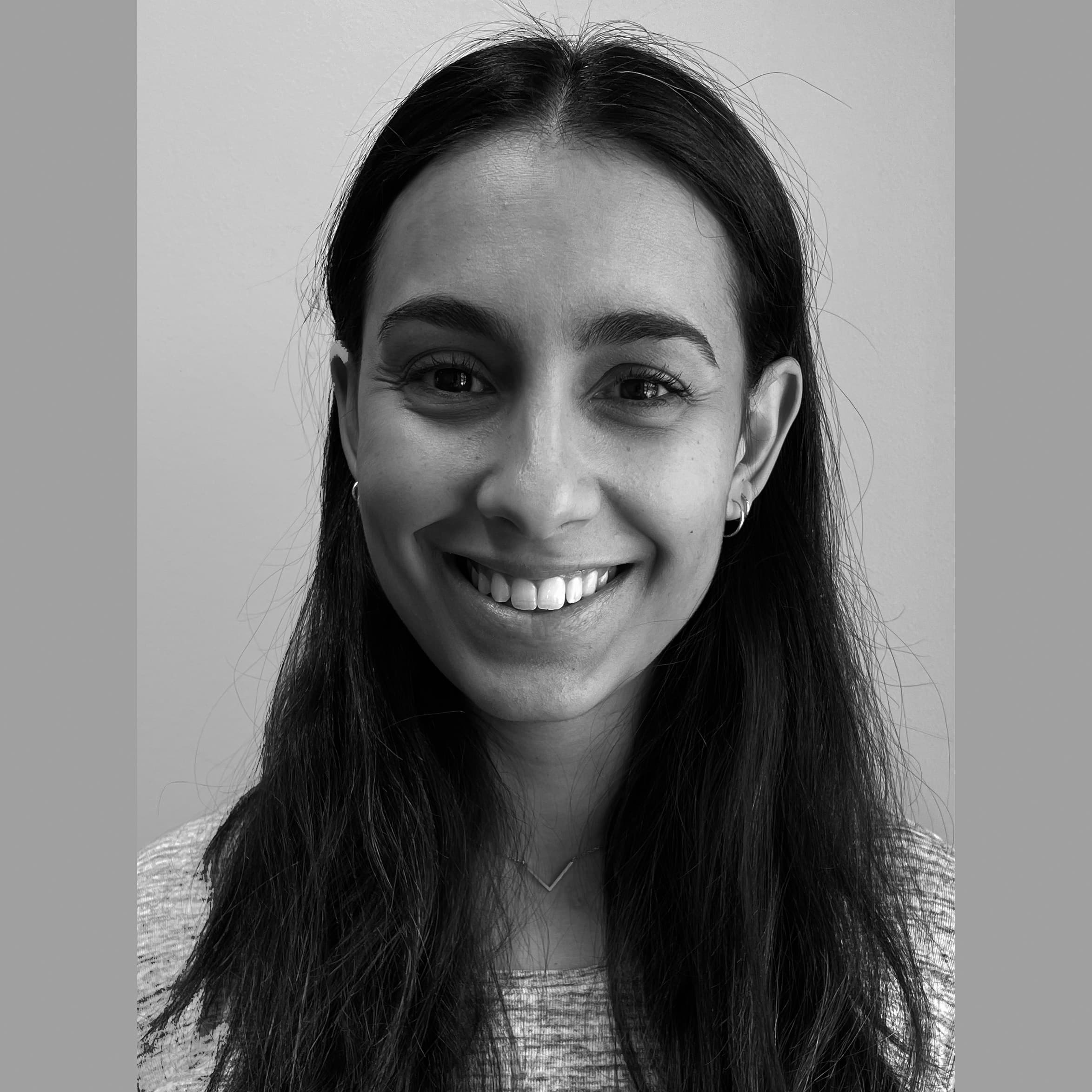 Svart-hvitt portrett av en kvinne med langt, mørkt hår, som smiler og ser rett inn i kameraet, med en ensfarget bakgrunn.
