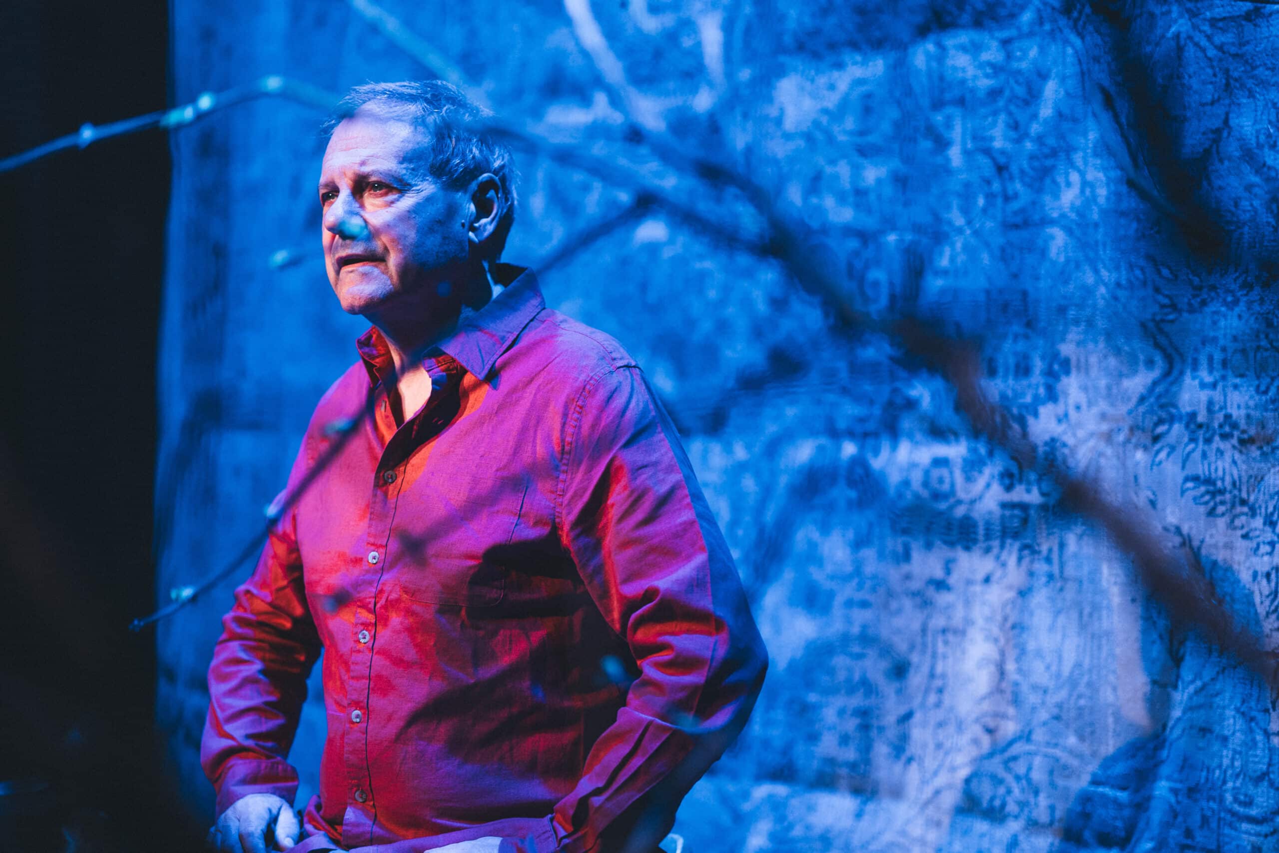 En eldre mann i rød skjorte står foran et mønstret blått bakteppe, opplyst av blått og rosa scenelys.