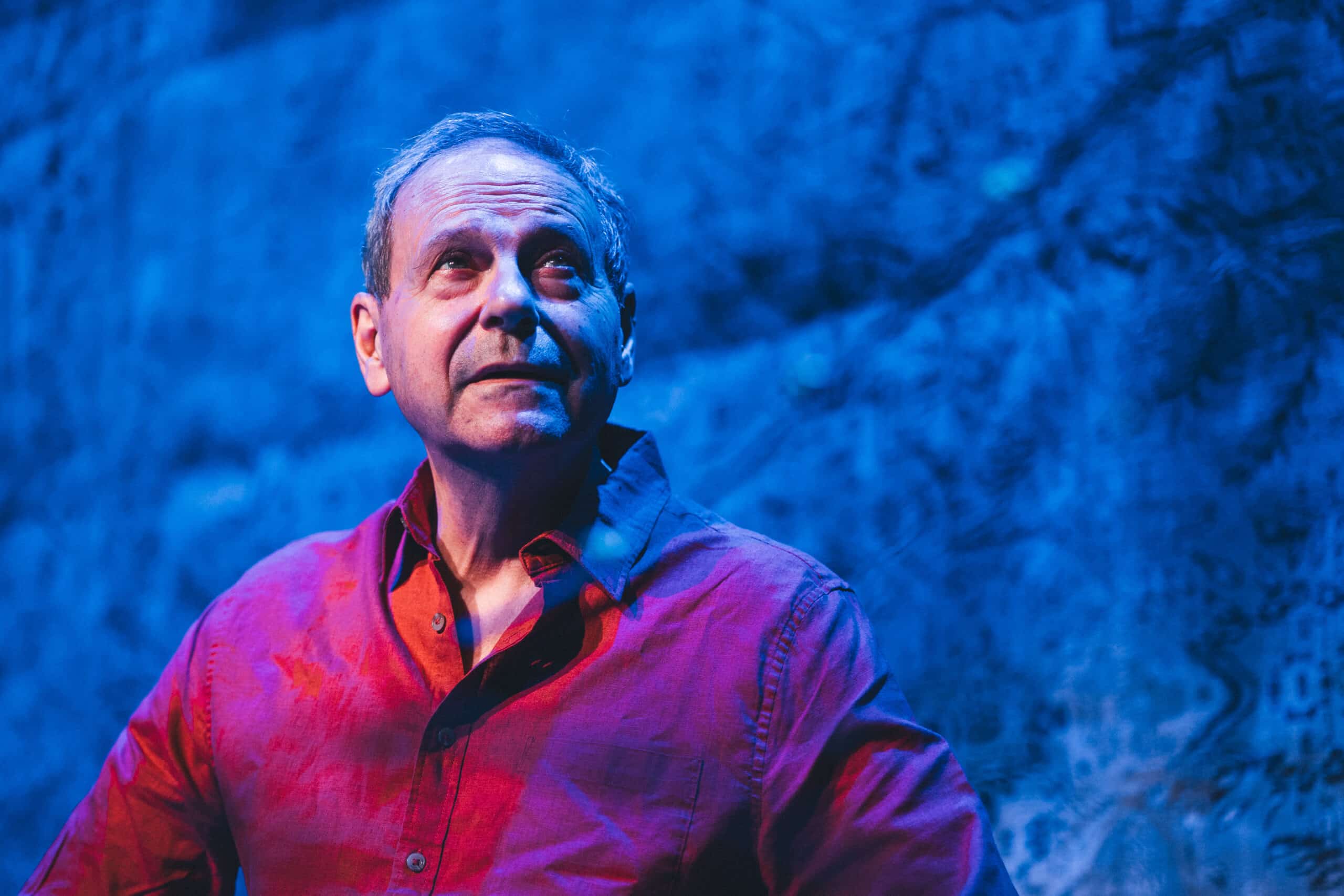 En mann i rød skjorte ser oppover i blått lys med en blå, strukturert bakgrunn bak ham.