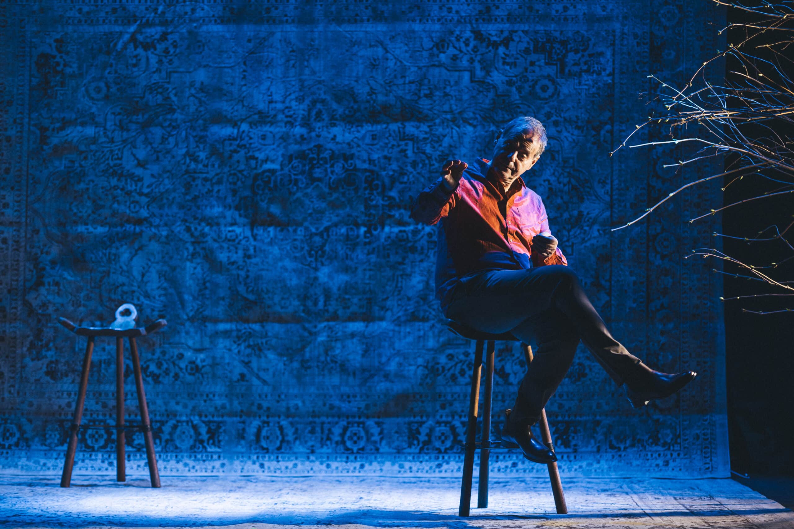 En mann i rød skjorte sitter på en krakk mot en blåmønstret bakgrunn og gestikulerer med hånden. En annen tom krakk med en gjenstand på står i nærheten.