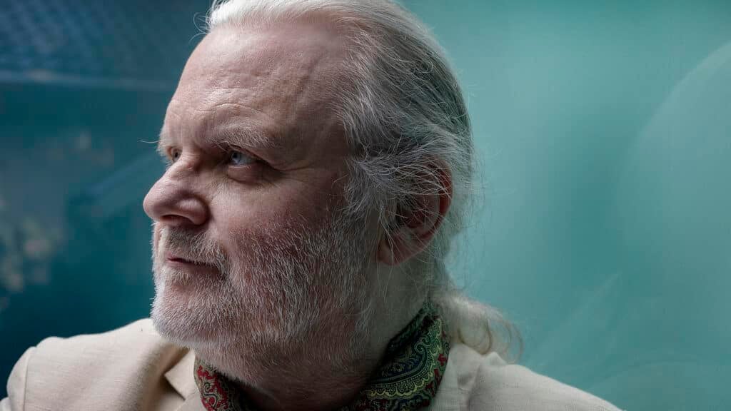 En eldre mann med hvitt hår og skjegg, kledd i lys dress og mønstret skjerf, ser til venstre mot en blågrønn bakgrunn.