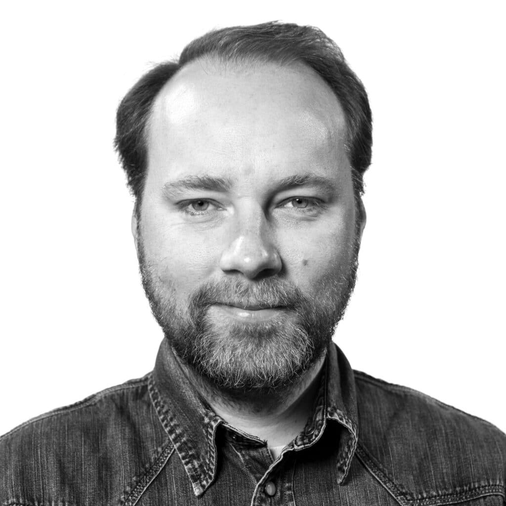 Mann med skjegg og bart iført jeansskjorte, vendt mot kamera og lett smilende, mot en vanlig hvit bakgrunn.