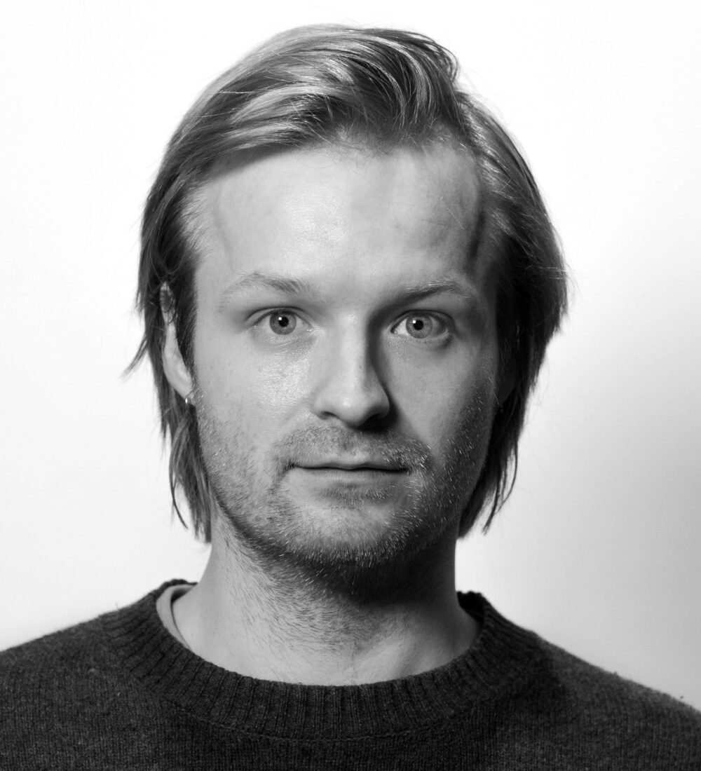 En mann med lyst hår og kort skjegg ser rett inn i kameraet mot en hvit bakgrunn. Han er iført en mørk genser.