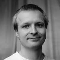 En mann med kort, lyst hår og et lite smil er avbildet i svart-hvitt, vendt mot kameraet og med nøytral bakgrunn.