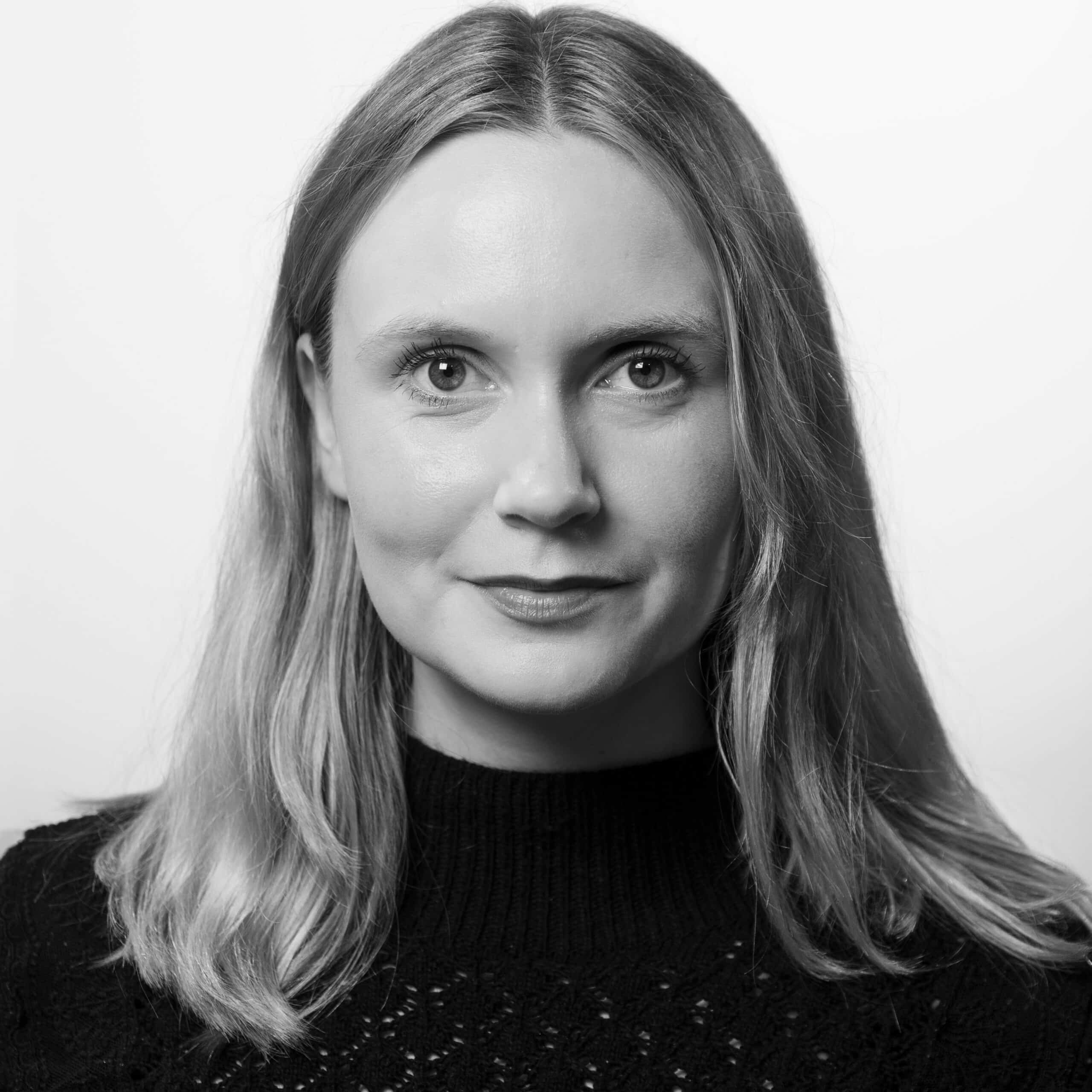 En kvinne med rett, skulderlangt hår og nøytralt uttrykk bærer en mørk, strukturert genser, fotografert mot en enkel, lys bakgrunn i svart og hvitt.