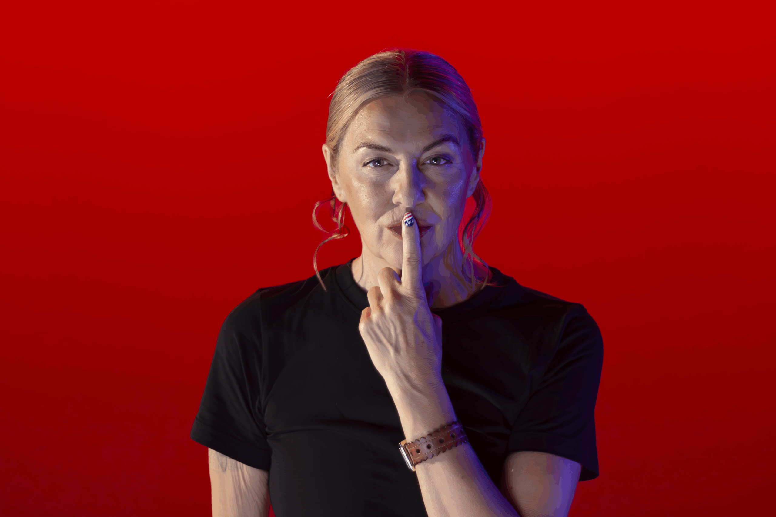 En kvinne i svart skjorte står mot en rød bakgrunn og holder pekefingeren mot leppene i en gest som antyder stillhet.