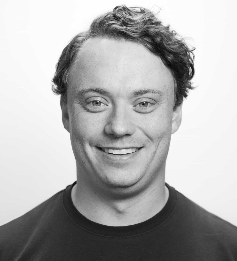Kent Andreas En mann med kort, bølget hår smiler til kameraet mot en ensfarget, lys bakgrunn. Han er iført en mørk skjorte med rund hals.