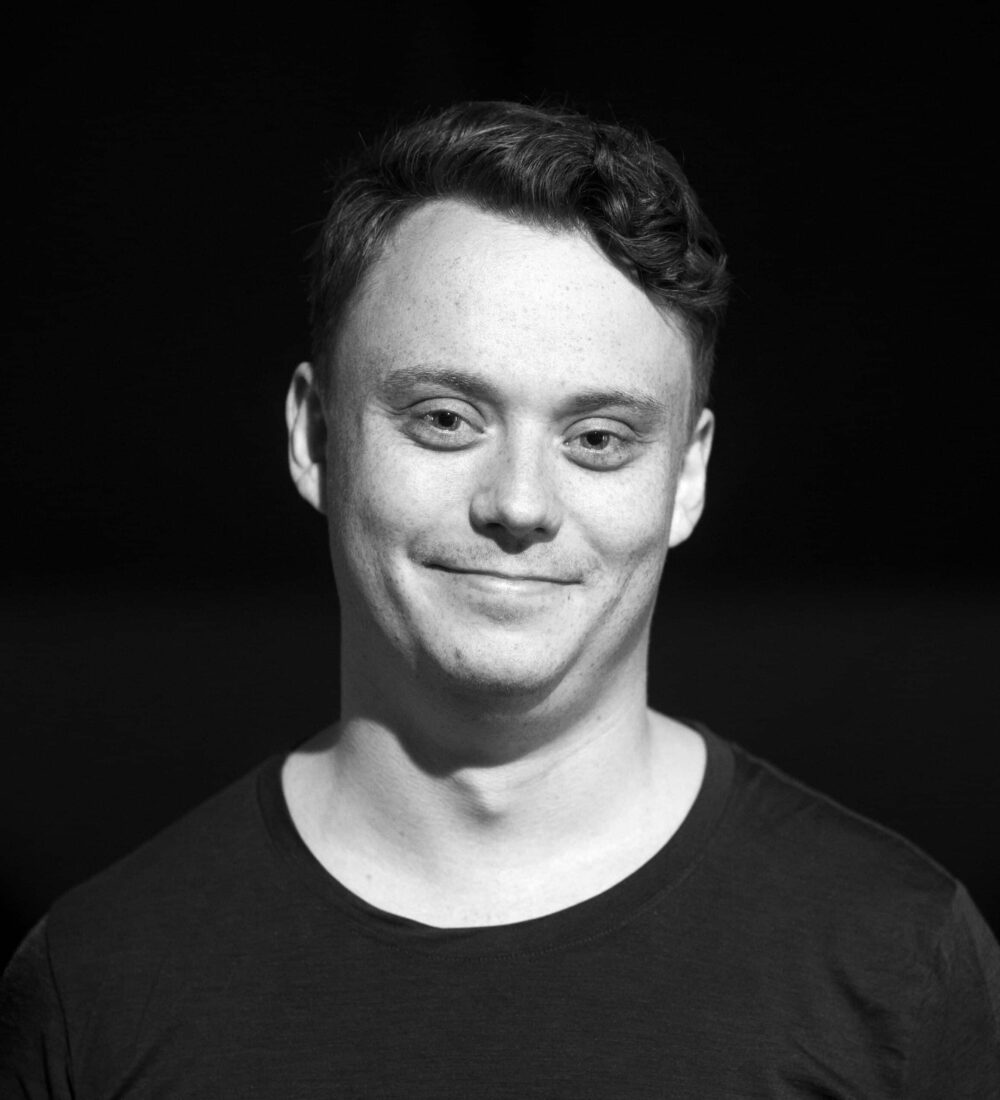 En person med kort, bølgete hår og en mørk T-skjorte står mot en mørk bakgrunn og vender mot kameraet med et lite smil.
