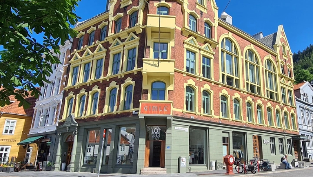 På et gatehjørne i en by står en treetasjes mursteinsbygning med gule kanter og store buede vinduer, med sykler parkert utenfor og et skilt med teksten "GIMLE".