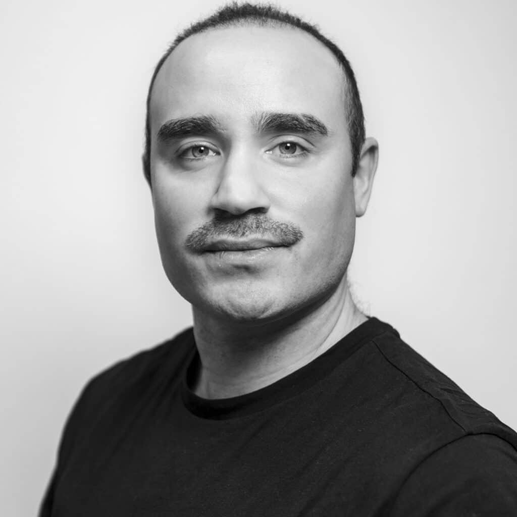 Svart-hvitt portrett av en skallet mann med bart iført svart skjorte.