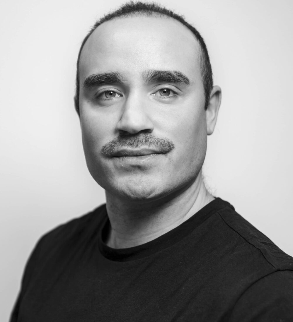 Kurt Svart-hvitt portrett av en skallet mann med bart iført svart skjorte.