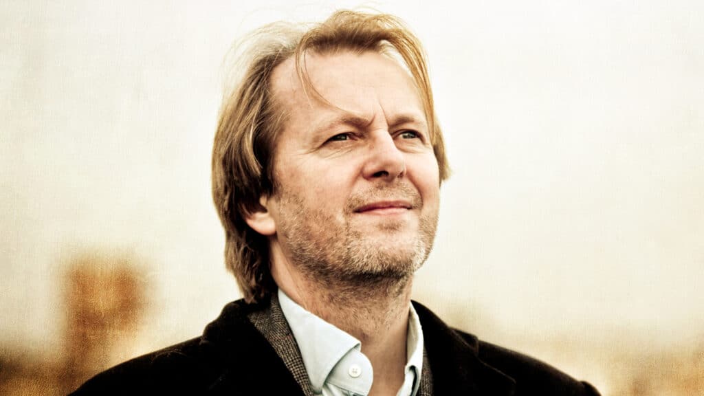 En middelaldrende mann med lysebrunt hår og kort skjegg ser litt oppover, iført skjorte med krage og mørk jakke, mot en nøytral, uskarp bakgrunn.