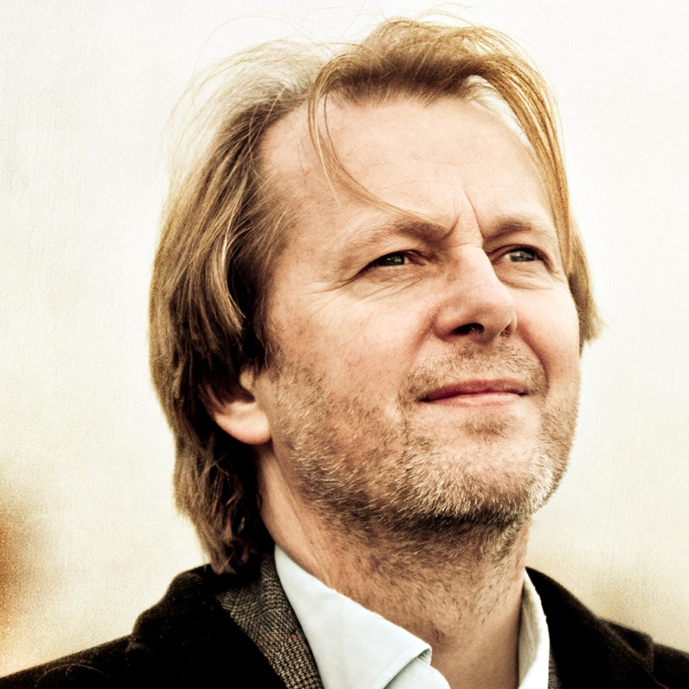 En middelaldrende mann med lysebrunt hår og skjeggstubber ser lett oppover med et nøytralt uttrykk, iført skjorte med krage og jakke, mot en uskarp, lys bakgrunn.