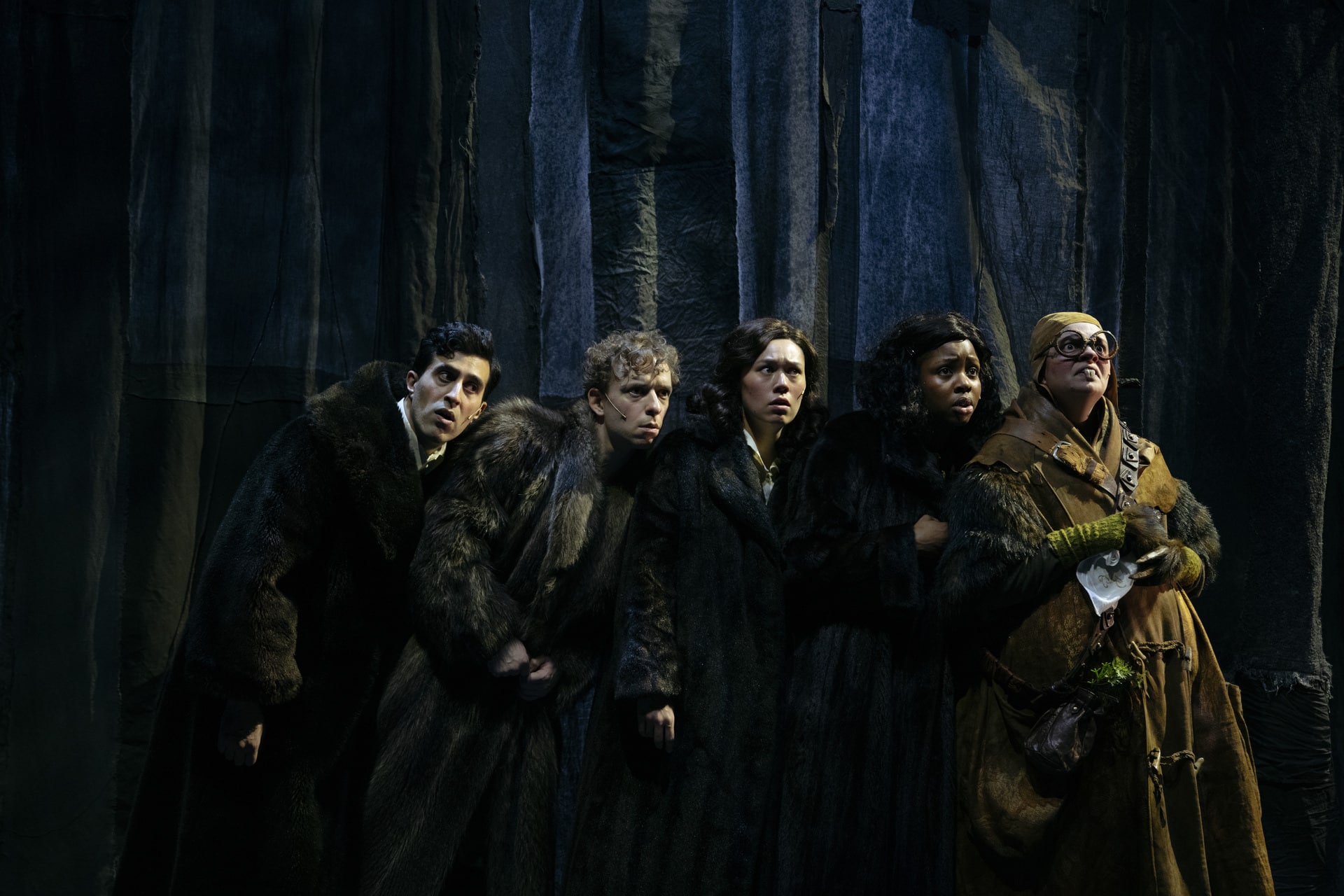 Fem skuespillere i mørke kostymer står tett sammen og ser intenst i samme retning, mot et bakteppe av mørkt, vertikalt stoff som ligner en skog.