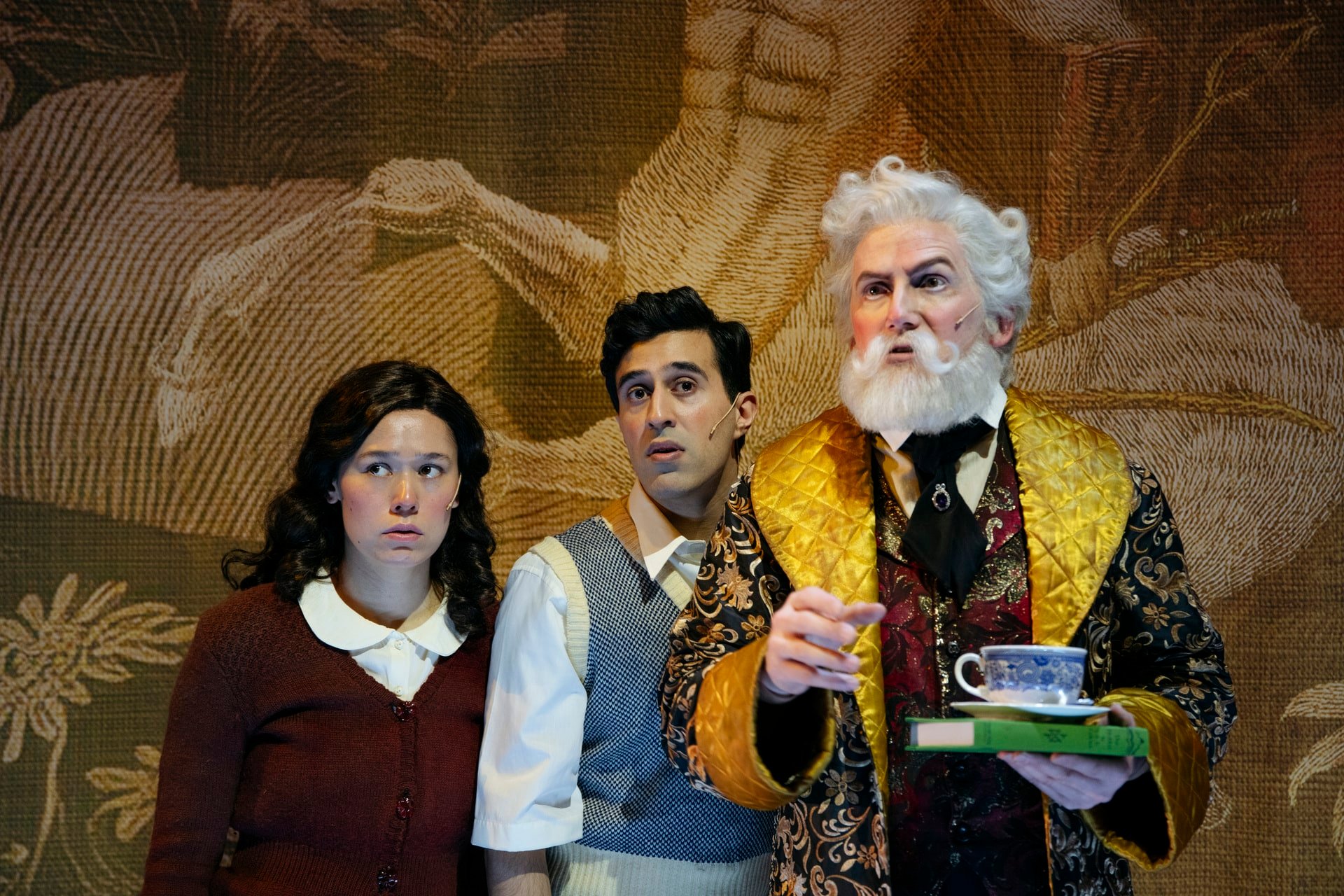 Tre personer står sammen innendørs; en kvinne og en mann ser bekymret ut, mens en eldre mann med hvitt hår og skjegg holder en tekopp og et fat, iført en utsmykket kappe.