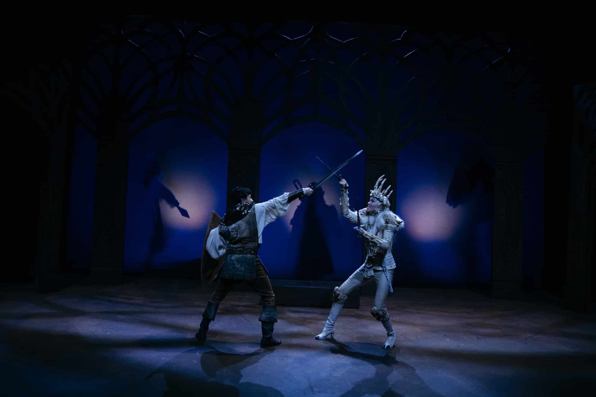 To skuespillere i forseggjorte kostymer utkjemper en dramatisk sverdkamp på en dunkelt opplyst scene med blå belysning og buede kulisser i bakgrunnen.