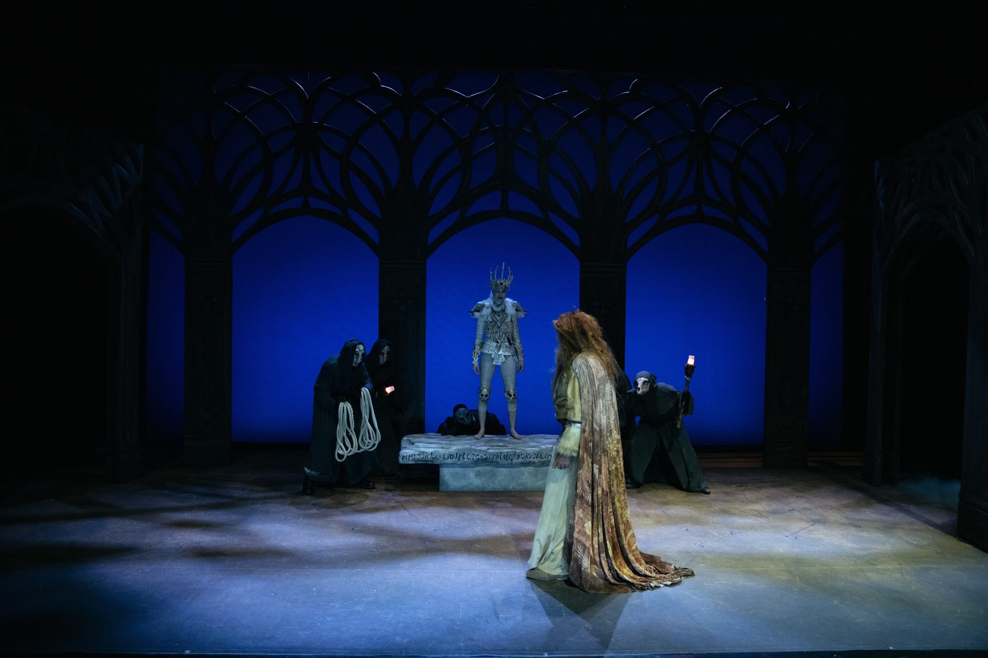 En scene med fem skuespillere: en i gevirdrakt står på en plattform, andre i mørke kapper og en i lang kappe holder lys, alt mot et blåbelyst bakteppe.