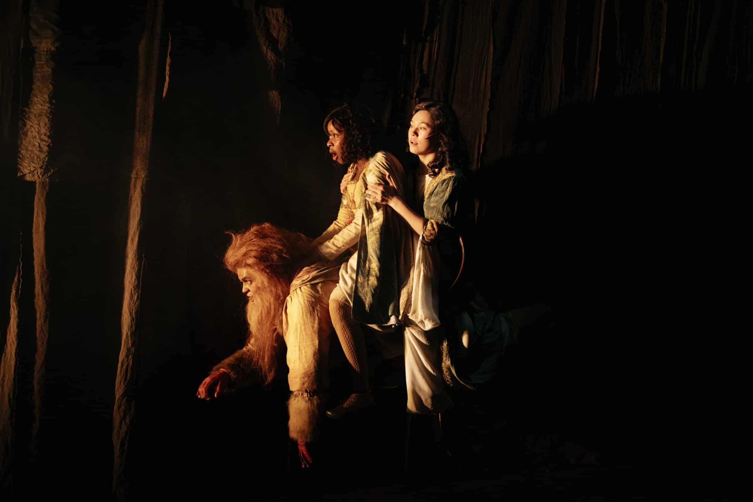 Tre personer i dramatisk belysning; en utkledd som løve sitter på alle fire, mens to andre i kostyme rir og går ved siden av ham i en dunkel, teatralsk setting.