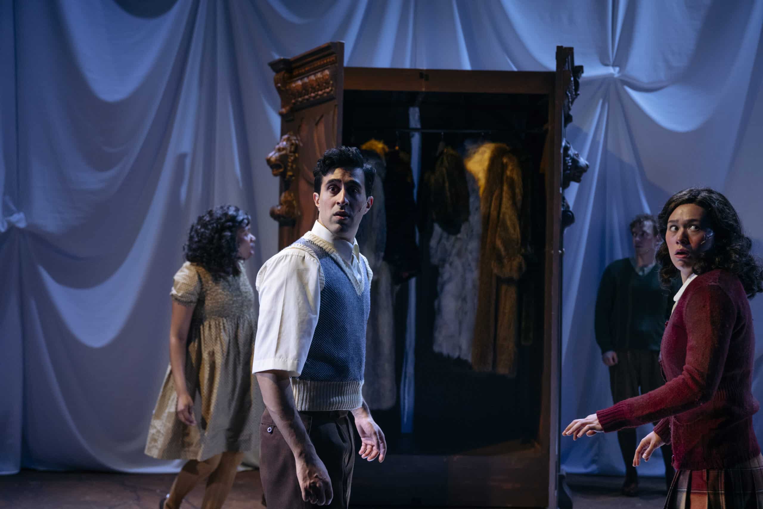 Fire skuespillere står forskrekket foran en åpen garderobe fylt med kåper; drapert hvitt stoff fungerer som bakteppe.