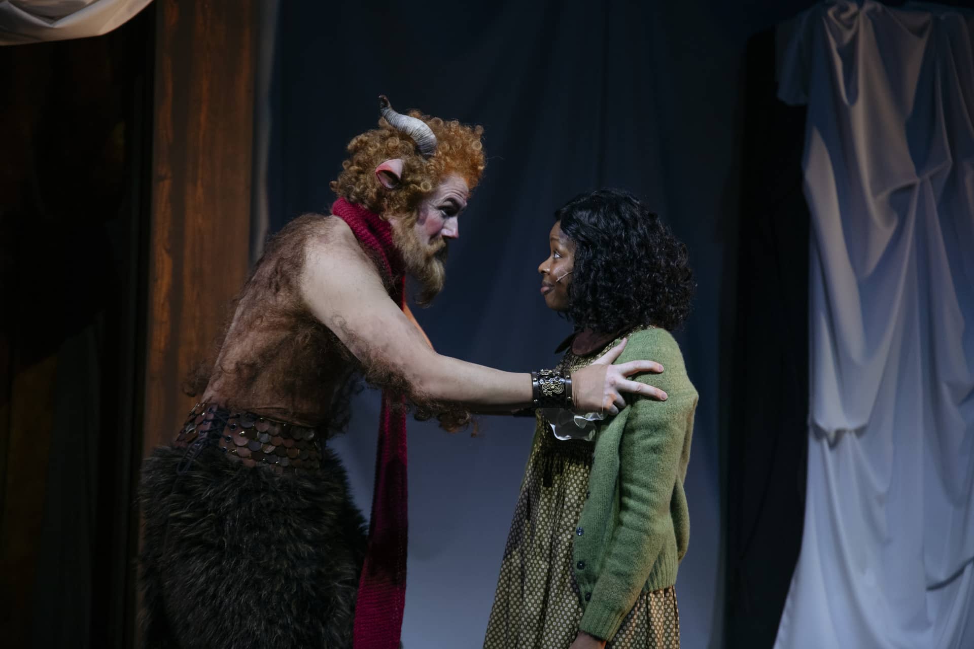 En person utkledd som en faun med horn og rødt skjerf snakker med en ung jente i grønn genser på en dunkelt opplyst scene.