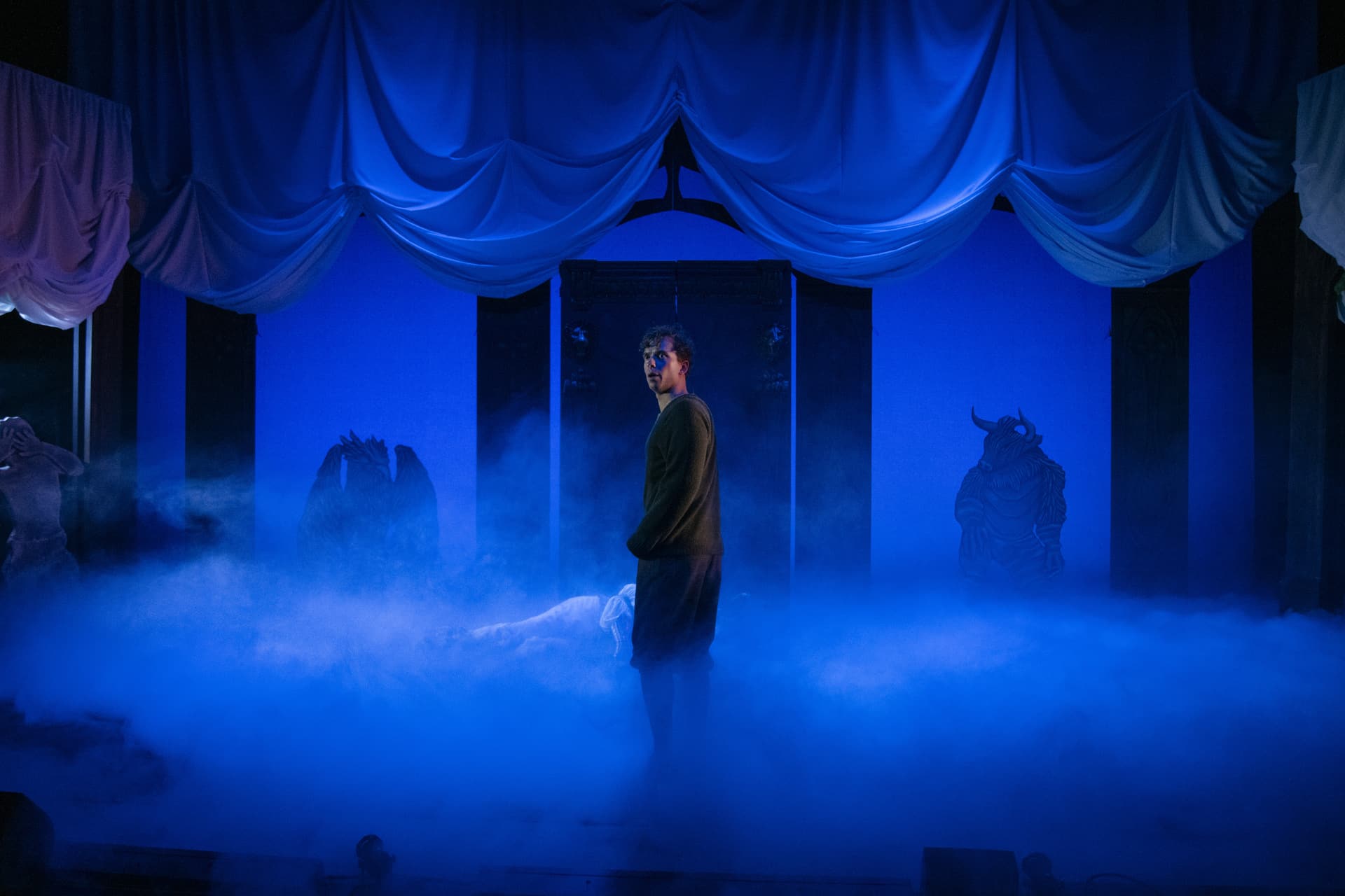 En person står midt på scenen i dramatisk blått lys i tåke, flankert av figurer i dyrekostymer mot en bakgrunn av drapert hvitt stoff.