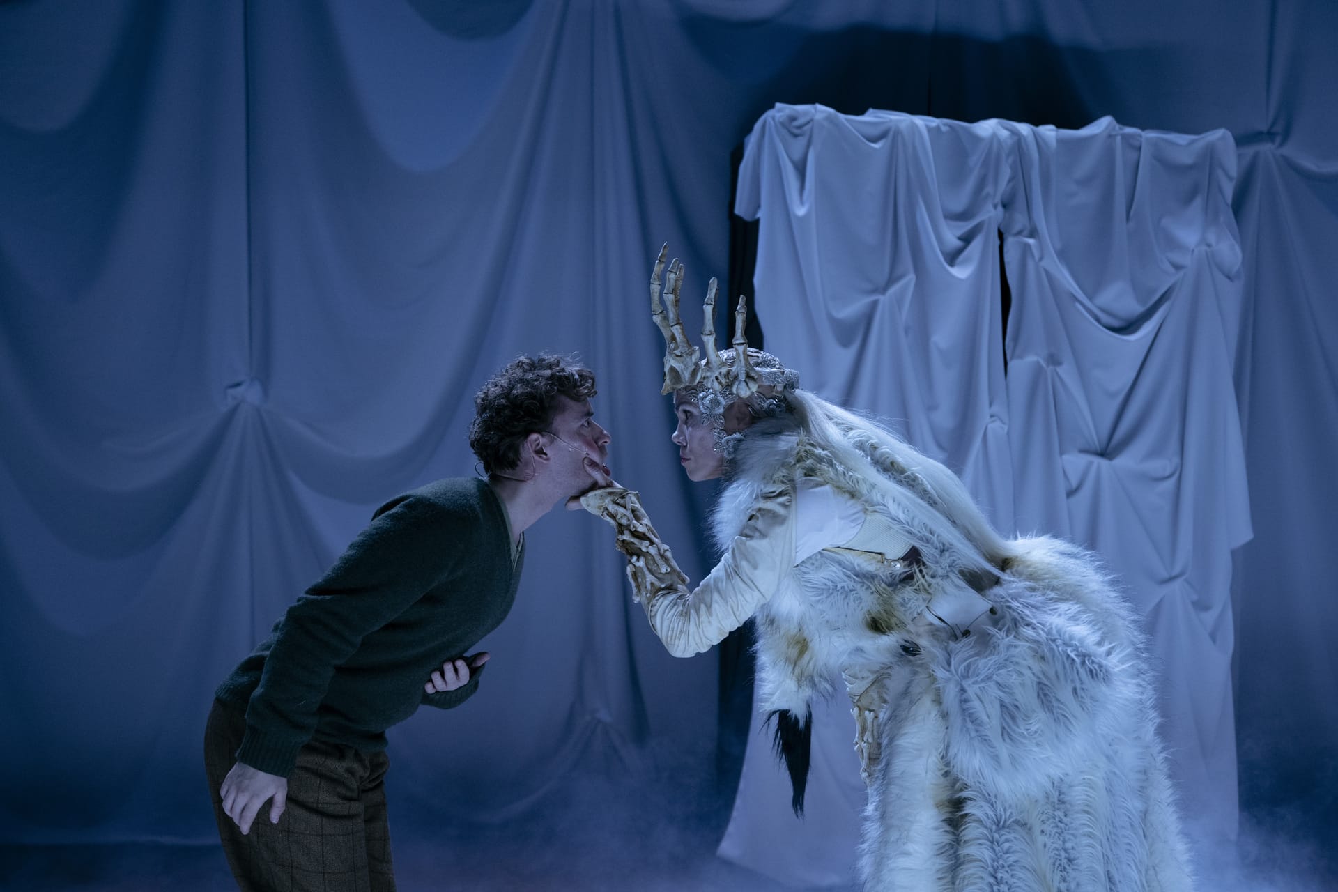 En person i grønn genser konfronteres med en annen person i hvit, pelskledd drakt med gevirhodepynt, mot et drapert hvitt stoff på scenen.