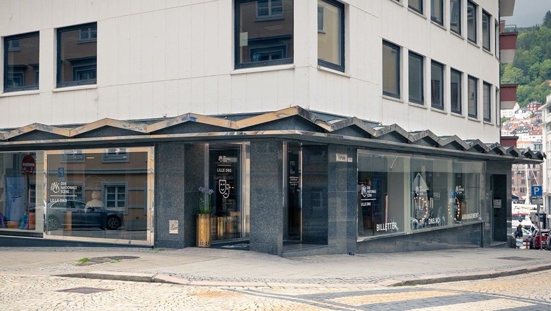 En hjørnebygning med store glassvinduer som viser forretningsskilt, beliggende i en brosteinsgate med litt grøntområder og parkerte biler i bakgrunnen.
