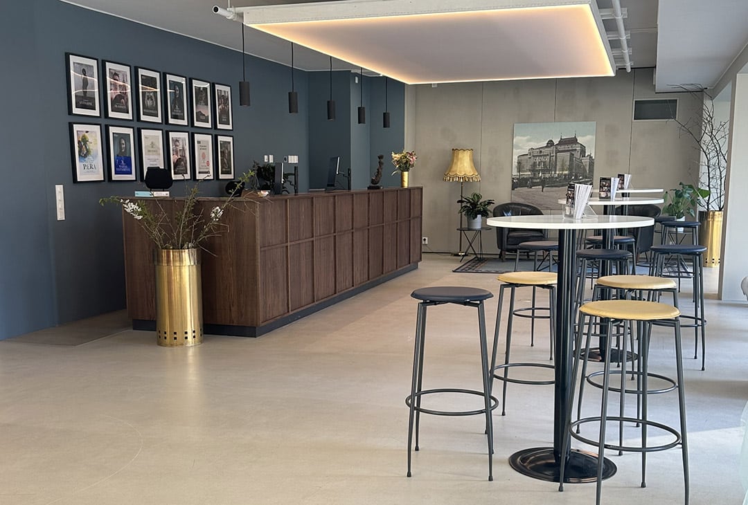 Moderne hotellobby med resepsjonsskranke i tre, innrammet kunst på en blå vegg, høye bord med barkrakker og en dekorativ gulllampe i hjørnet.