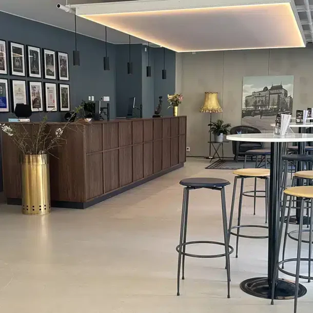 En moderne hotellobby med en resepsjonsdisk i tre, veggkunst, høye bord med krakker, planter og dempet belysning.