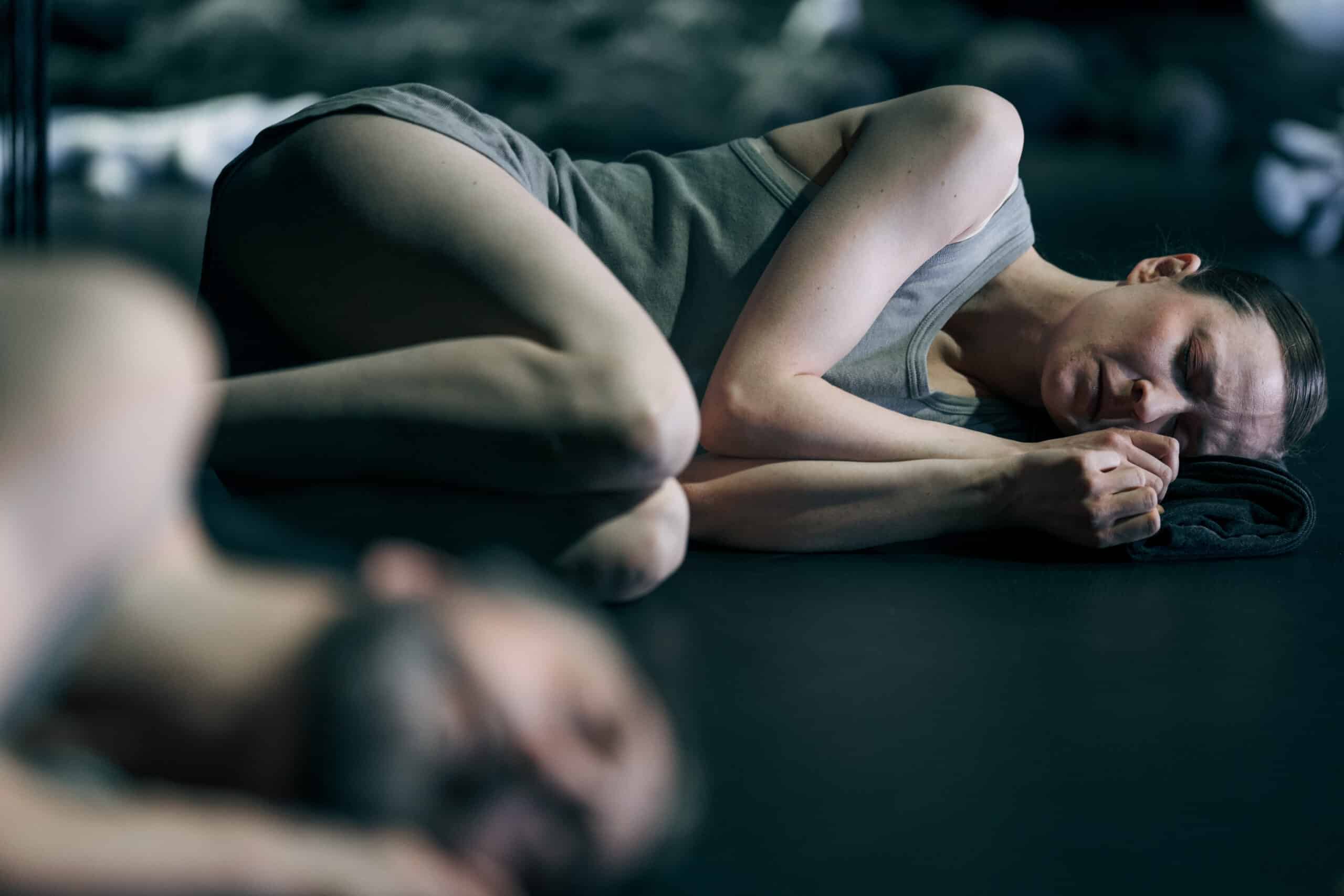 To personer som ligger på et mørkt gulv, den ene i fokus og den andre uskarp. Personen i fokus er krøllet sammen, iført en ermeløs skjorte. Scenen er svakt opplyst.