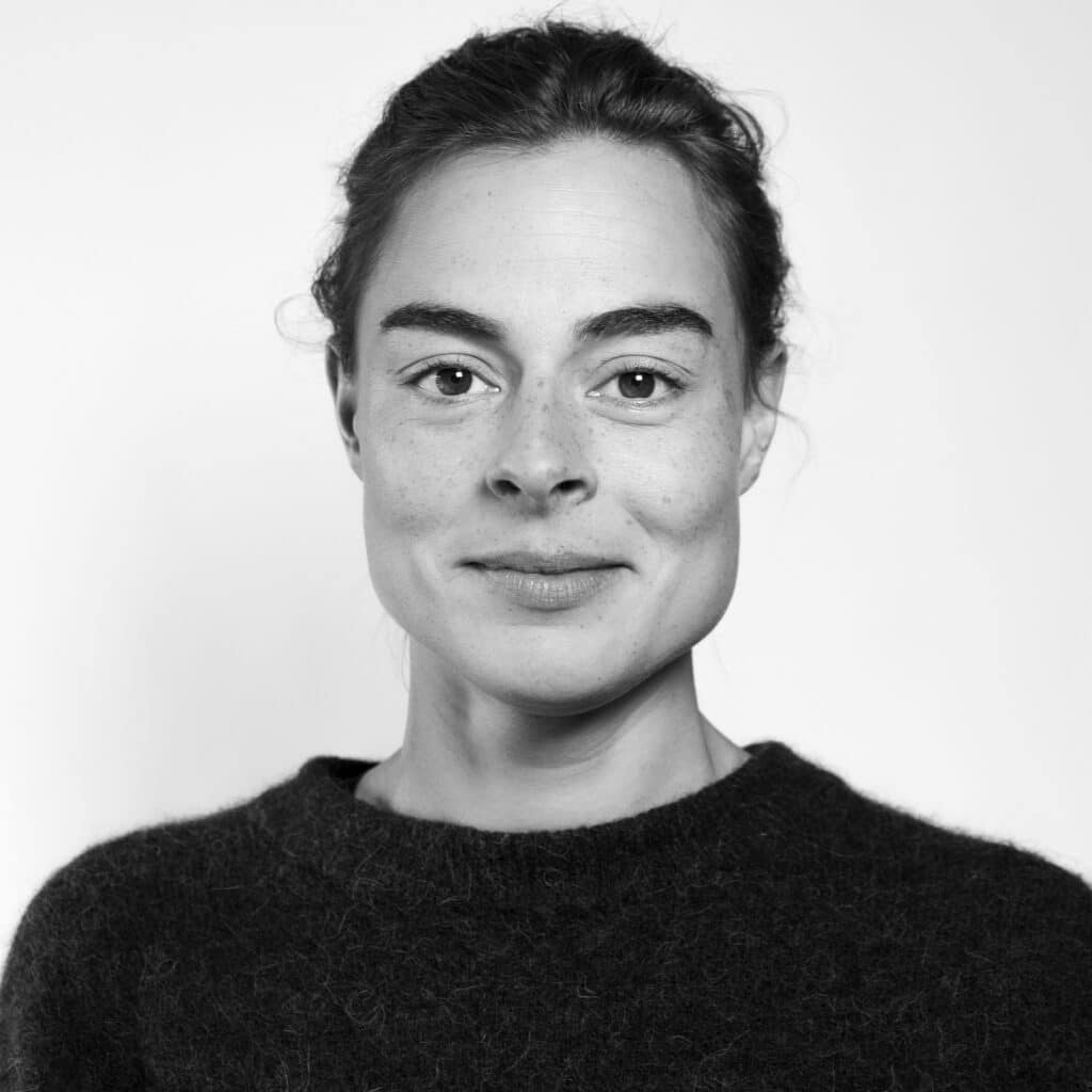 En kvinne med mørkt hår og mørk genser ser direkte inn i kameraet mot en enkel, lys bakgrunn.