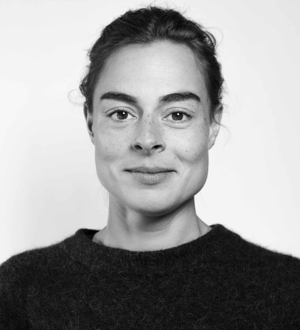 En kvinne med mørkt hår og mørk genser ser direkte inn i kameraet mot en enkel, lys bakgrunn.