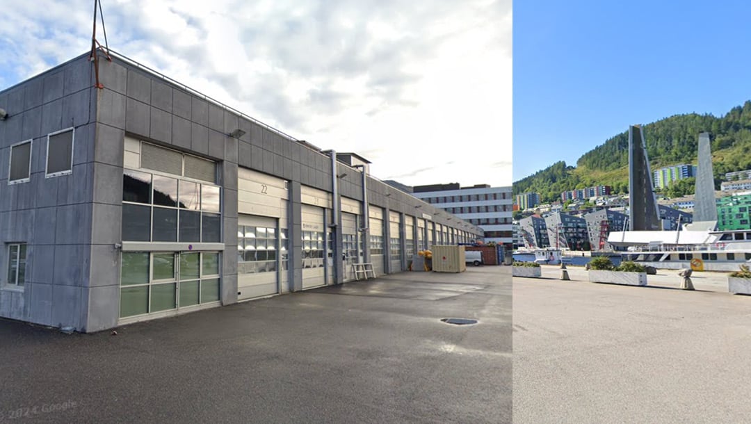 En grå industribygning med store garasjeporter ligger ved siden av en asfaltert tomt, med moderne bygninger og grønne åser i bakgrunnen.